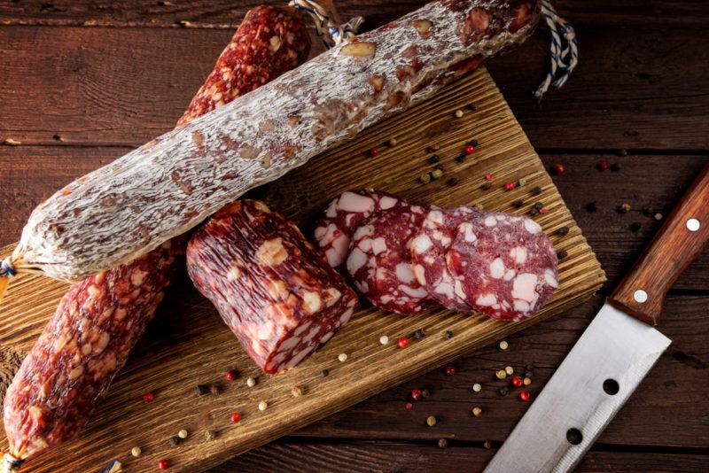 A wooden board with various types of fresh salami, peppercorns and a knife