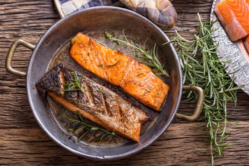 A steel fry pan with two cooked salmon fillets, one the right side up and the other upside down