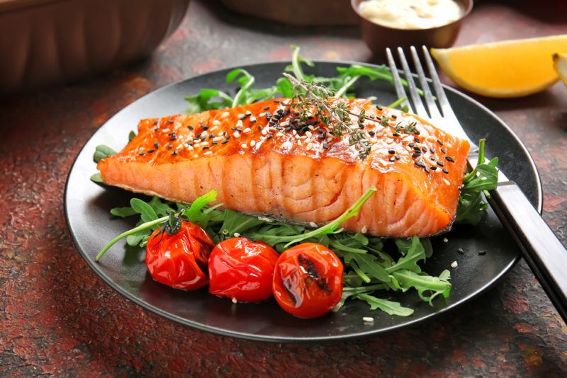 A gray plate with cooked salmon, greens, and tomatoes, and a fork