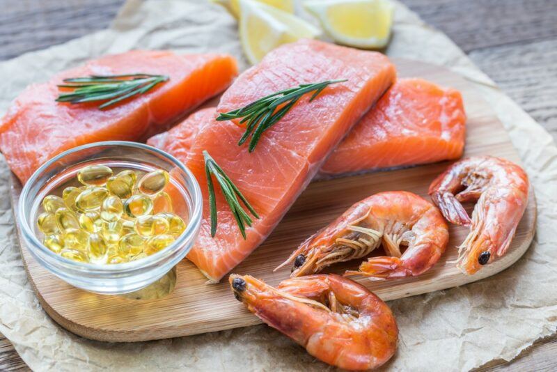 A wooden board with pieces of raw salmon, prawns, and a small glass bowl of fish oil