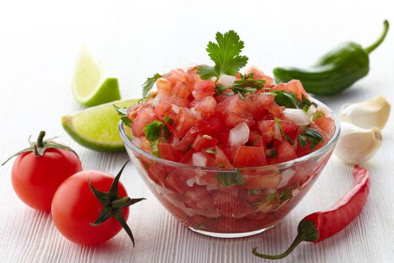 A glass bowl of salsa with tomatoes and limes on a table