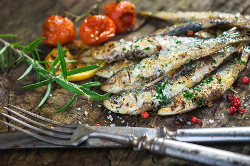 A selection of sardines cooked on a board with a knife and fork, tomoatoes and rosemary