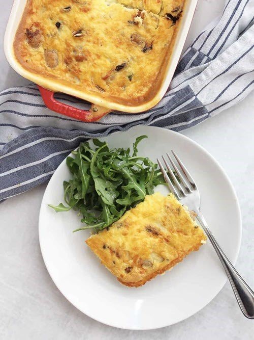 A white plate with a piece of sausage and egg breakfast casserole, with a larger dish of the casserole in the background