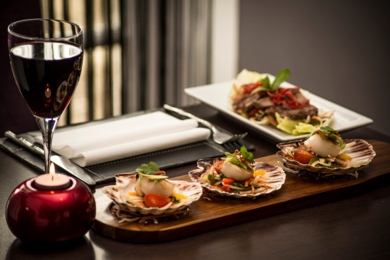 A wooden tray with various fresh scallops, next to a glass of red wine and another tray of food