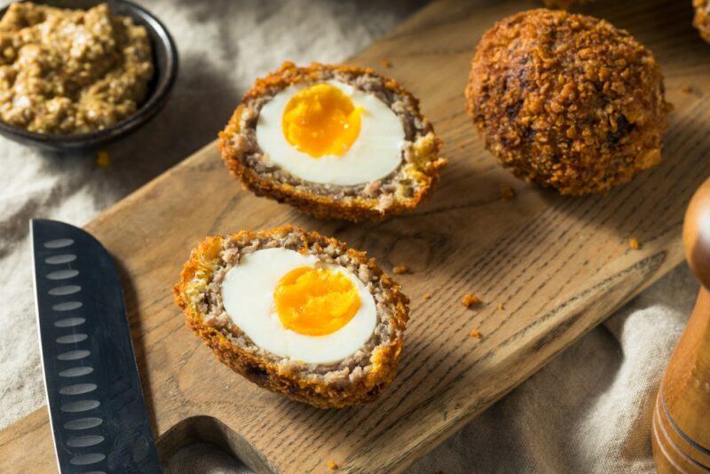 A wooden board with one Scotch egg that has been cut in half and another whole one, with a knife at the end of the board