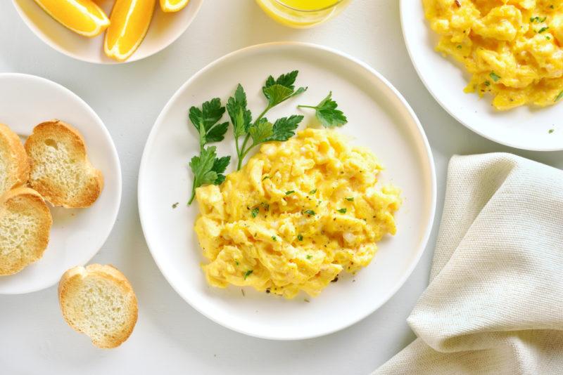 A whilte plate of scrambled eggs with some greens, with another plate of eggs nearby and one of bread