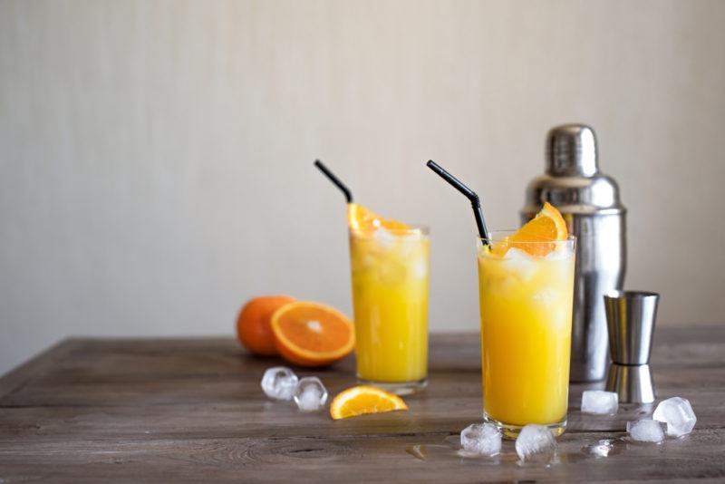 Two screwdriver cocktails with straws next to ice, oranges, a cocktail shaker and a jigger