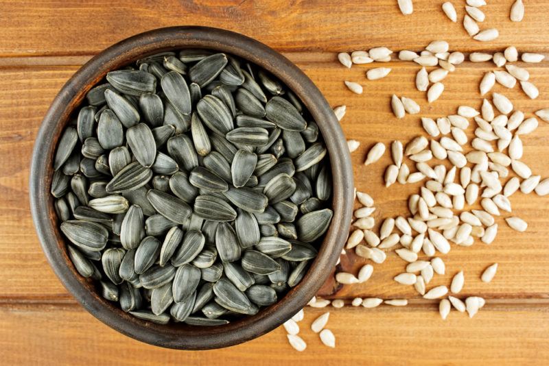 A bowl of sunflower seeds on a table with shelled seeds next to it