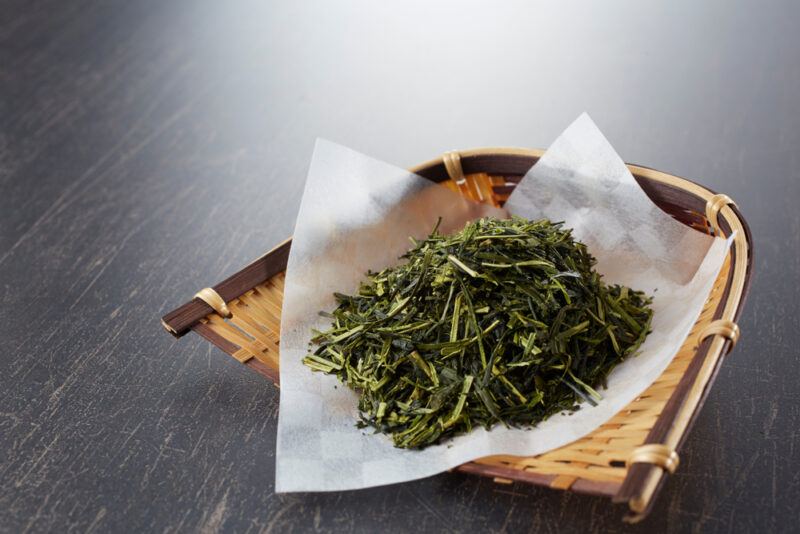 on a black wooden surface is a weaved basket with a white baking paper on it with loose Sencha tea