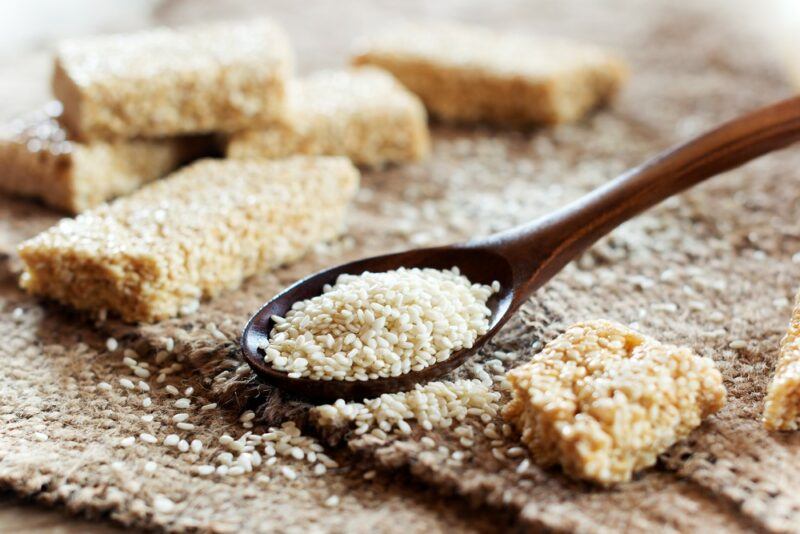 A collection of sesame treats on a table with a wooden spoon that contains more sesame seeds, plus some seeds scattered on the table