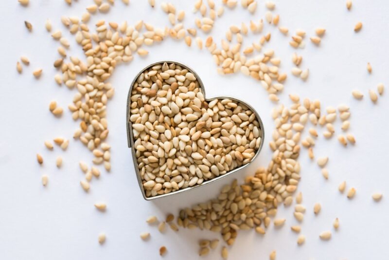 A heart shaped container with sesame seeds, with more seeds scattered across the table