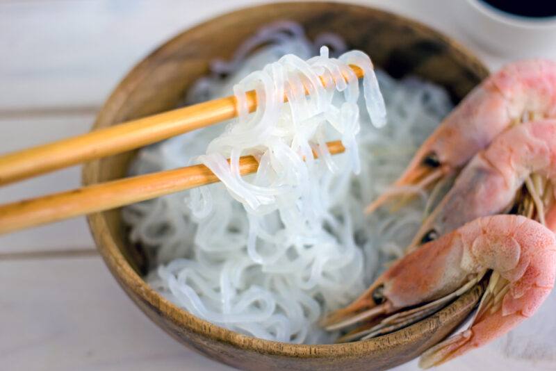 a wooden bowl with Shirataki noodles with three shrimps beautifully placed at its right-hand rim with a couple of wooden chopsticks opposite it