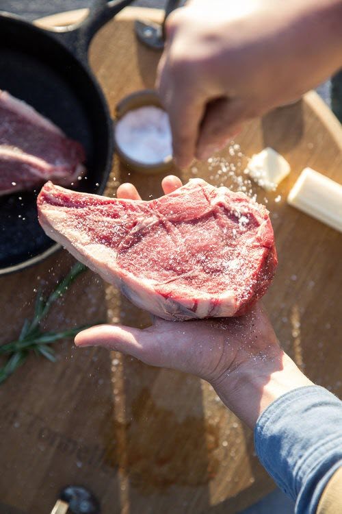 A hand holding a raw t-bone steak and sprinkling salt.