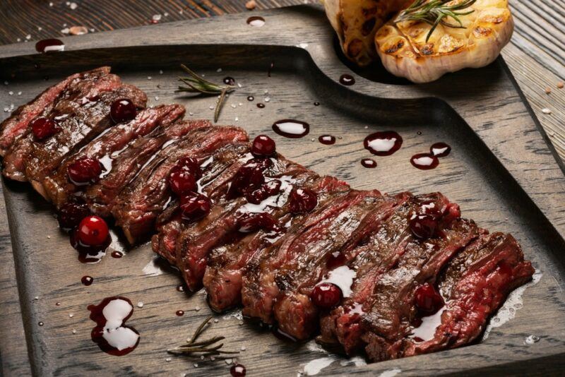 A large wooden board with a cooked and sliced piece of skirt steak, next to roasted garlic