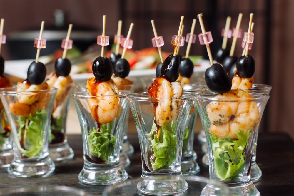 Various shot glasses with skewers that contain lettuce, shrimp, and an olive