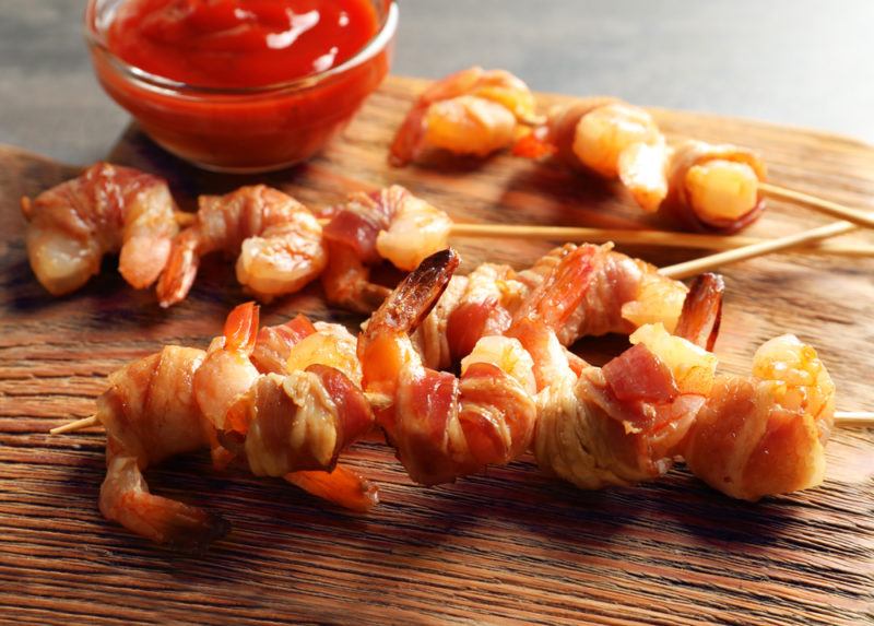 A wooden tray with skewers of shrimps wrapped in bacon next to a glass container of dip