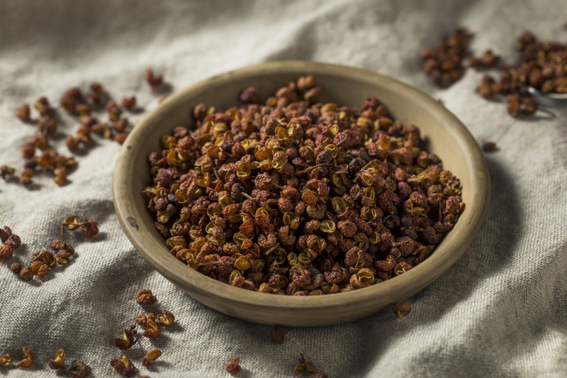 Light brown colored ceramic bowl full of sichuan peppers laid out on a khaki brown cloth with loose sichuan peppercorns around it.