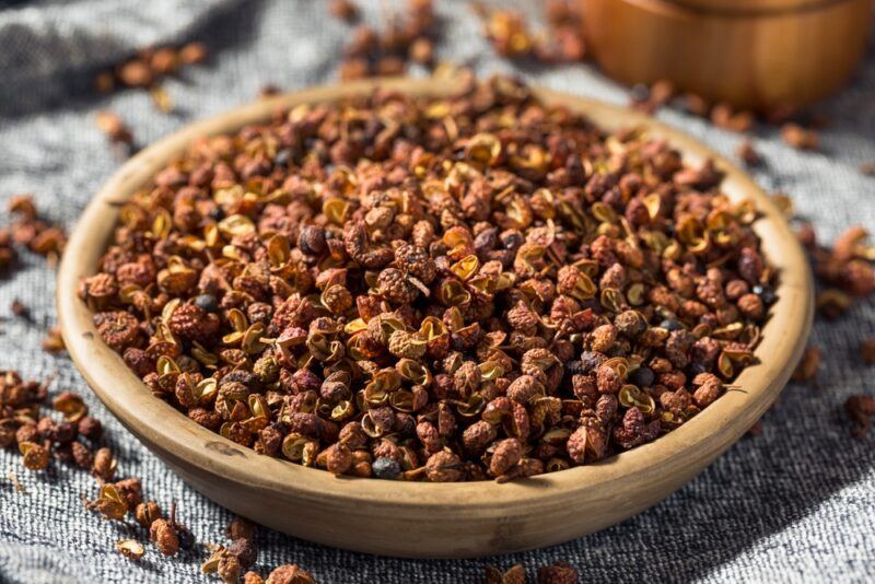 a closeup image of a wooden bowl full of Sichuan peppercorns resting on top of a grey table cloth with loose Sichuan peppercorns