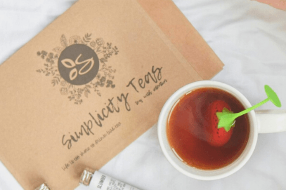 Simplicity tea box in background and a white ceramic tea cup with a strawberry shaped and colored diffuser in a cup of steaming  tea on a white back drop 