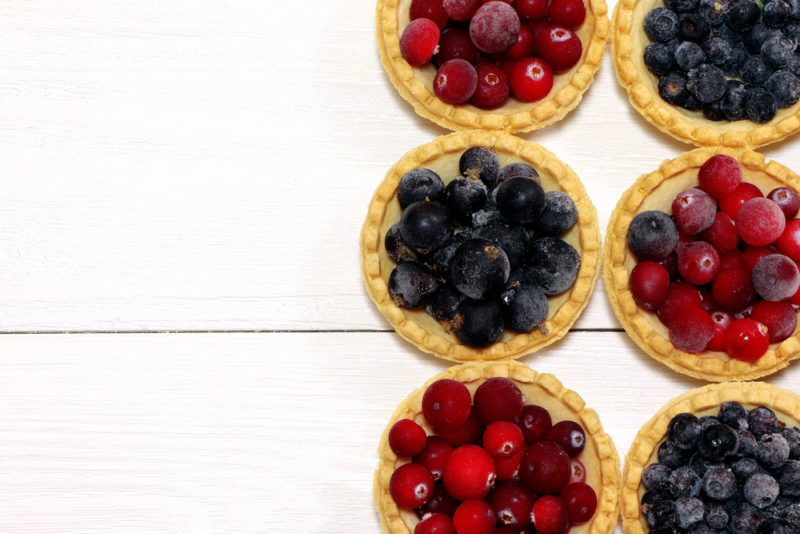 Six fruit tarts on a white table, three with red berries and three with black berries