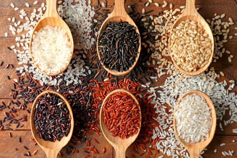 Six spoons on a table, each containing a different type of rice with the same types of rice under the spoons as well