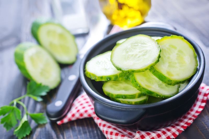 This photo shows a small black bowl filled with cucumber slices sitting on a red and white checked cloth near a knife and two more cucumber slices and a sprig of parsley on a wooden table.