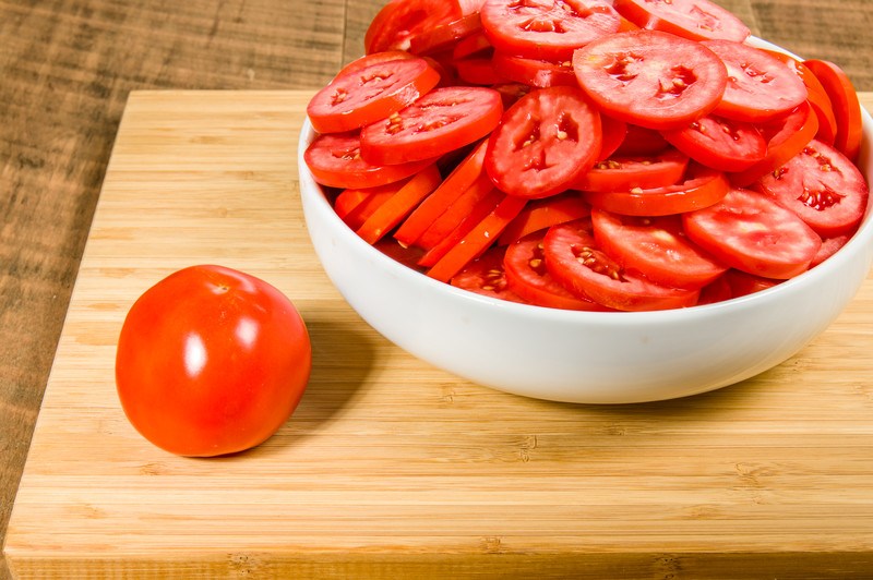 This photo shows white bowl filled with sliced tomatoes sitting on a wooden cutting board near a whole tomato, with the cutting board itself lying on a wooden table.