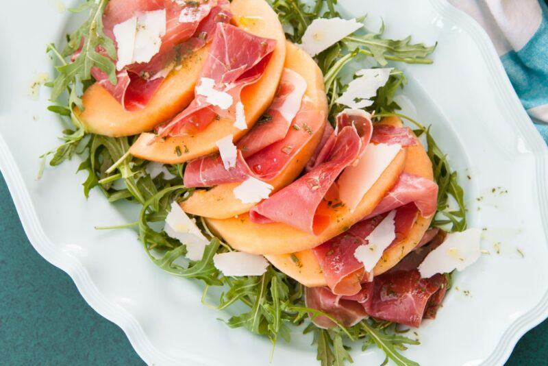 Slices of cantaloupe on a white table with greens, ham, and cheese, as part of an entree