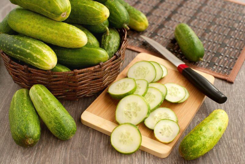 A basket of whole cucumbers, next to a wooden board with sliced cucumbers, and a few whole cucumbers on the table