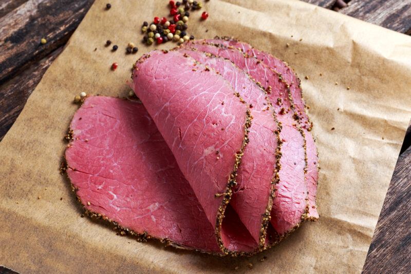 A piece of paper with sliced pastrami and some peppercorns