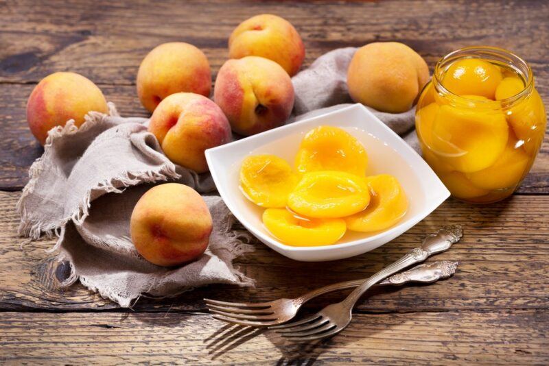 A wooden table with fresh peaches and a white dish of peaches from a can