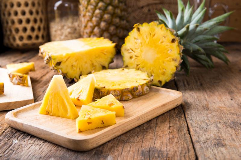 A wooden board with sliced pineapple, with the rest of the pineapple in the background