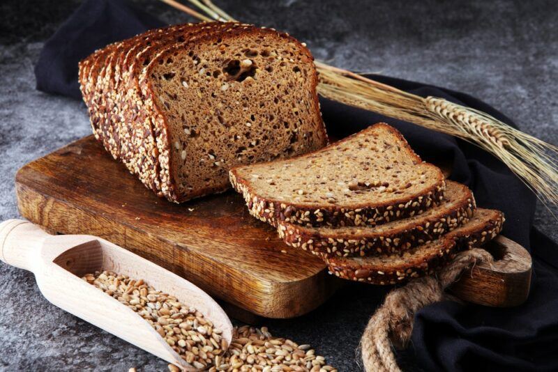 A wooden board with a loaf of rye bread and some slices, next to a scoop of the grains