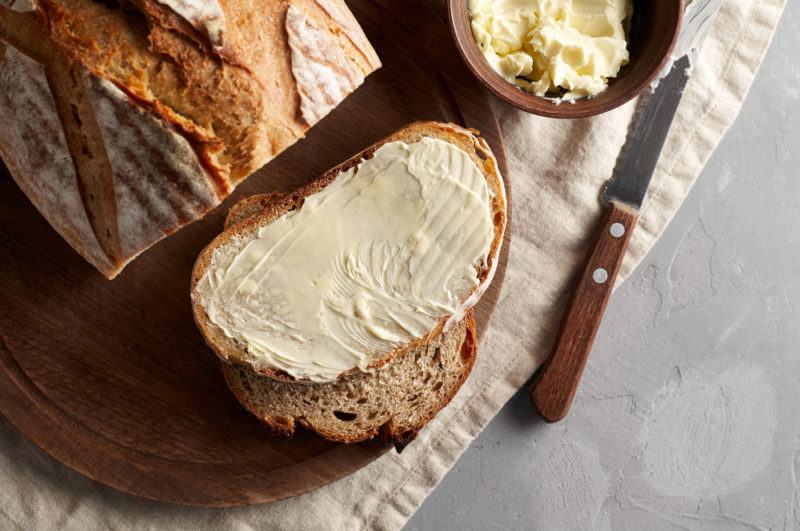 A loaf of rye bread where a piece has been sliced and spread with butter. The knife and a bowl of butter are visible in the image too.