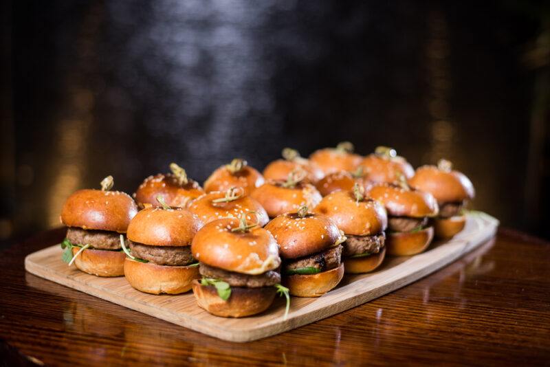 A wooden board, resting on a wooden surface, holds 15 hamburger sliders against a dark background.