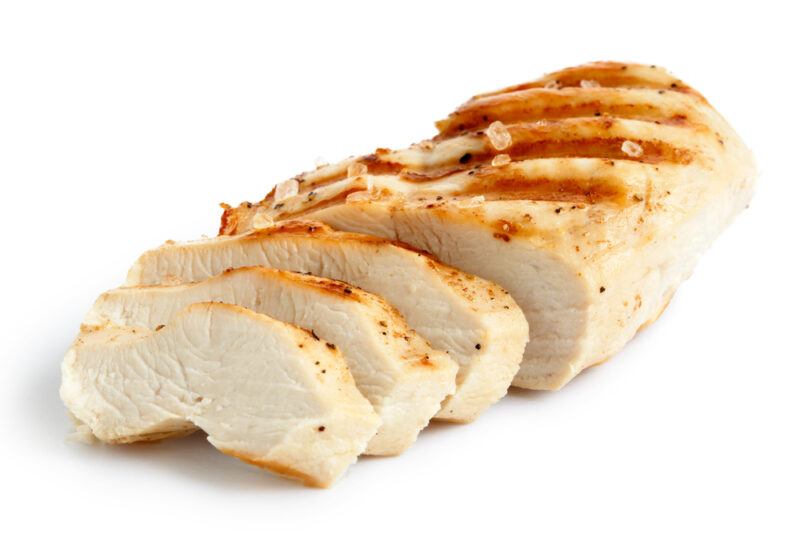 A piece of grilled chicken breast with three slices cut off the end of it rests against a white background.