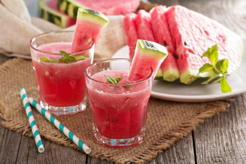 A wooden table with a cloth, two glasses of watermelon slushie and a plate of watermelon slices