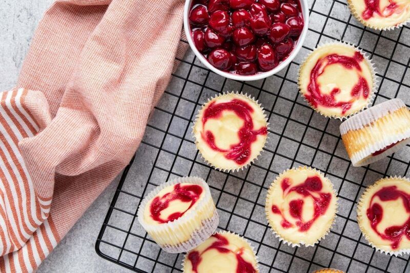 A wire tray containing a selection of small berry and vanilla cheesecakes, next to a pink cloth and a bowl of berries