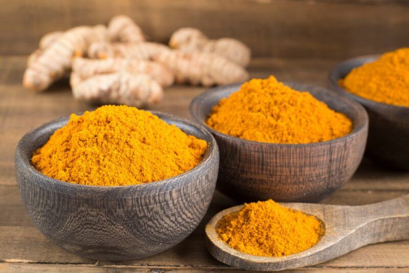 Three containers of turmeric powder next to a spoon of the powder and some turmeric roots in the background