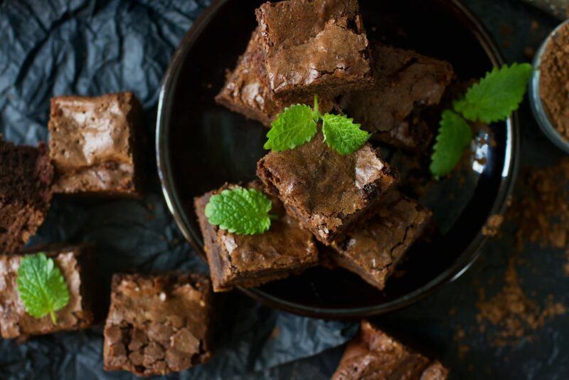 A dark bowl containing chocolate brownies, with more brownies on a dark background