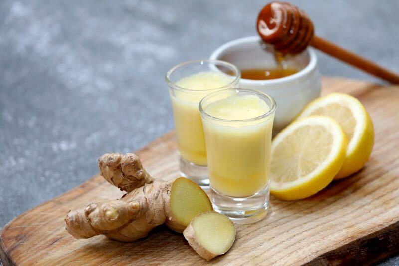 Two small ginger shots on a wooden board with ginger, lemon slices, and honey