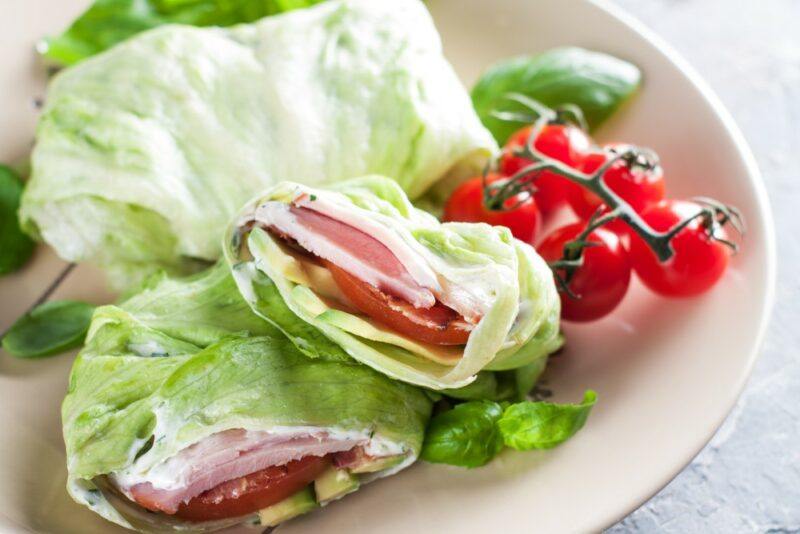 A white plate with wraps made using lettuce, meat, and cheese, next to more lettuce and some tomatoes