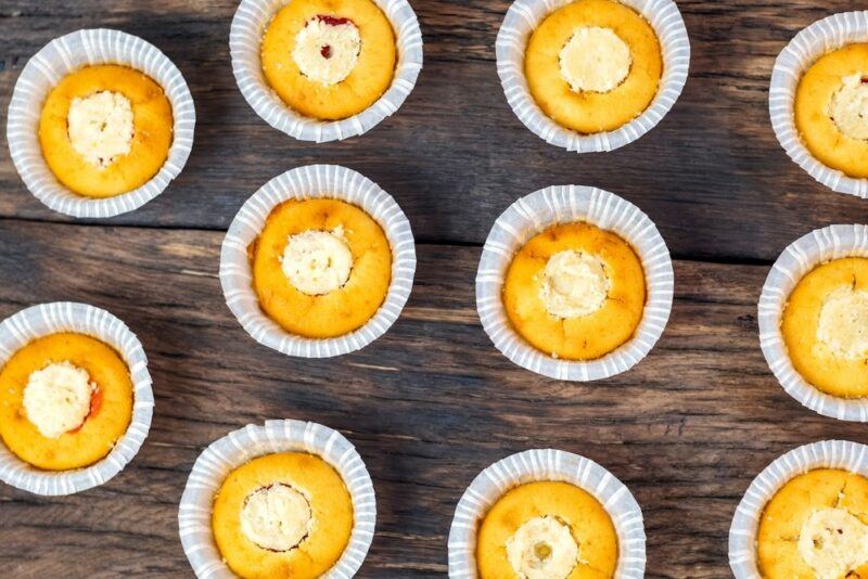 A wooden table with many small muffins. They all have their tops removed so they can be stuffed with a cheesecake filling