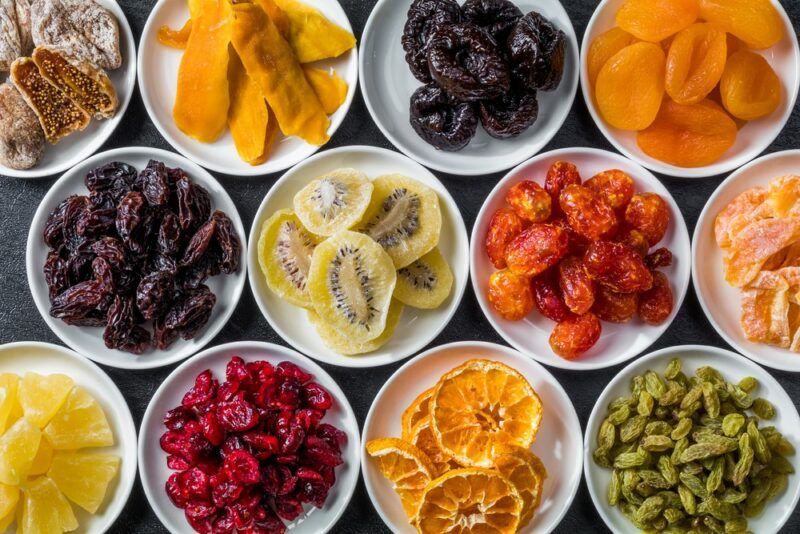 Small white dishes containing different types of dried fruit, like cranberries, oranges, apricots, kiwifruit, figs, and dates.