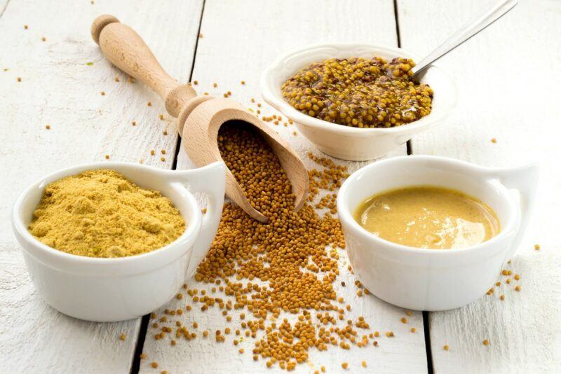 Three dishes containing different types of mustard, with a wooden scoop containing mustard seeds in the middle