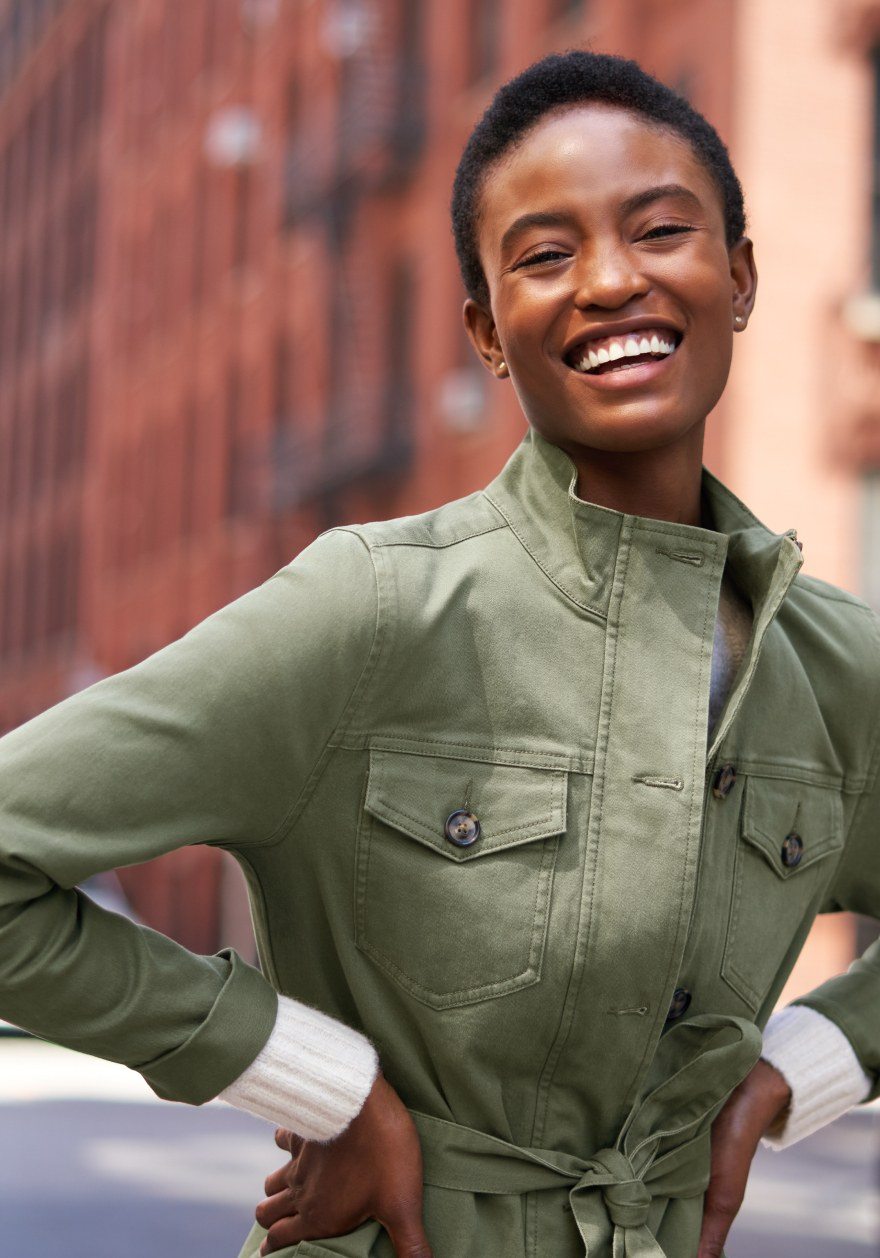 Smiling woman standing outside on a city block in a casual brushed denim sage green coat with the white cuffs of her sweater peeking out at the wrist