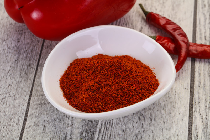 A white bowl with smoked paprika on a wooden table with fresh and dried pepper at the back.