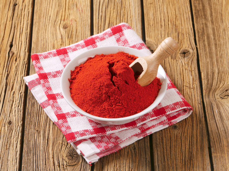 White ceramic bowl full of smoked paprika with wooden scoop, resting on a white and red checkered table napkin on a wooden table.