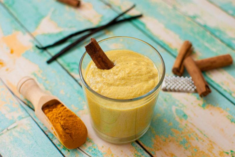 A turmeric smoothie on a light blue table, with turmeric, vanilla and cinnamon around the glass