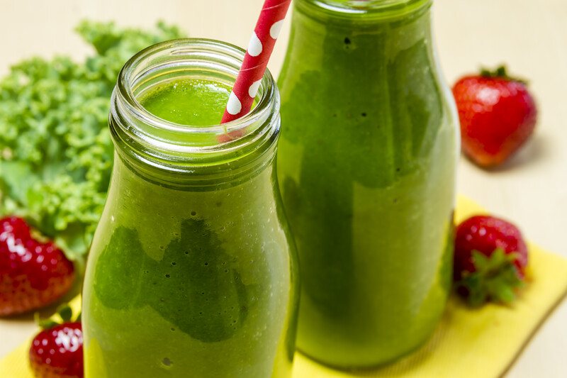 This photo shows two glass bottles filled with green smoothies on a yellow cutting board with strawberries and kale and a beige background.